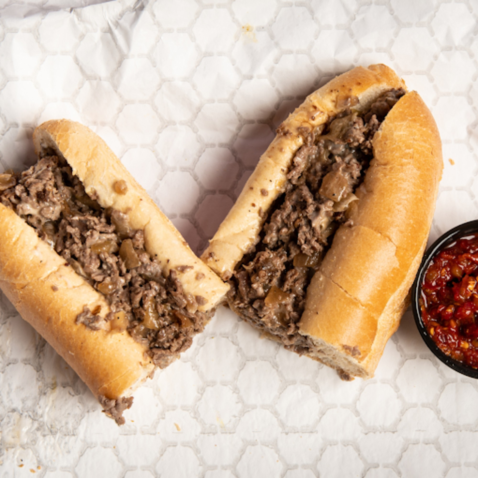Two halves of a Franzone's Pizzeria cheesesteak sandwich with a side of red sauce.